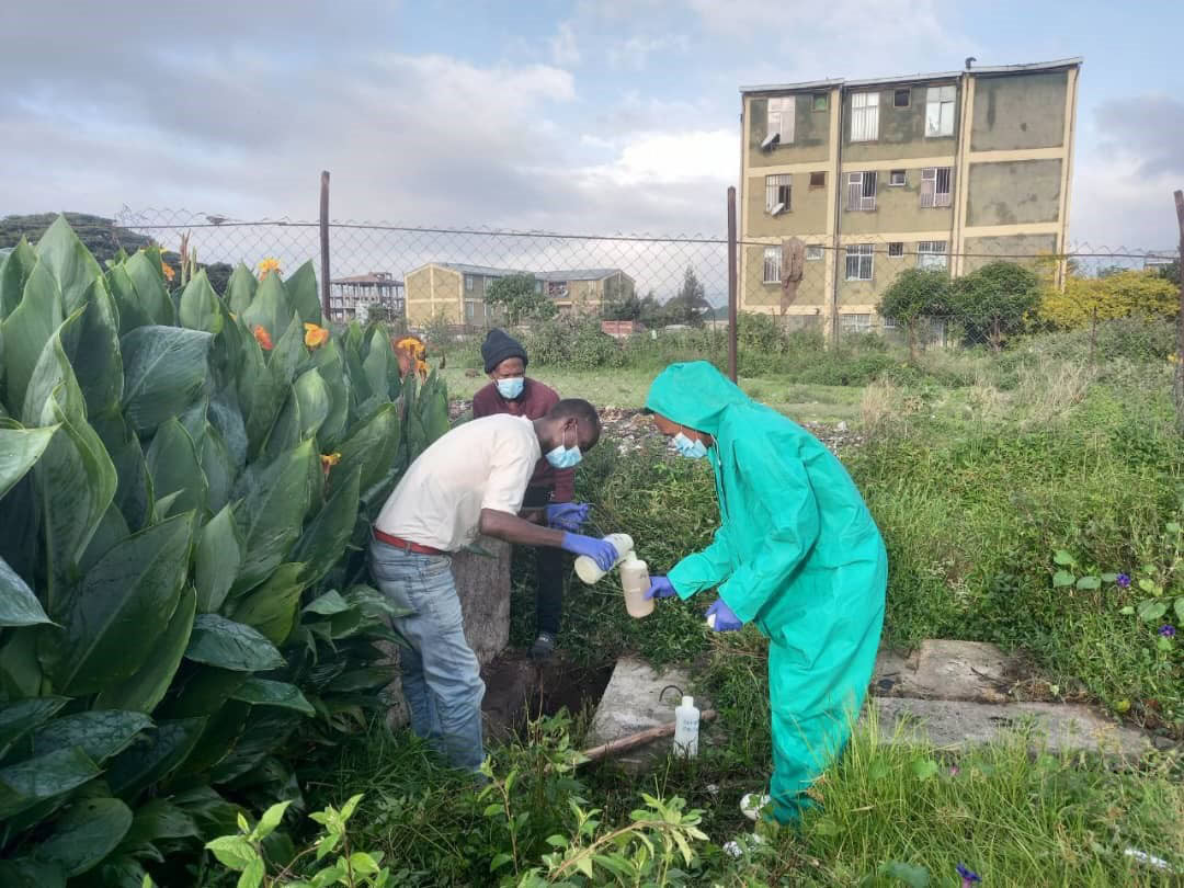 Three men in protective gear pouring water samples between bottles surrounded by greenery and apartment block in the background