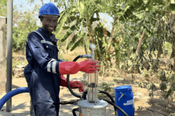 Man with blue overalls, red gloves and a blue hardhat stood smiling as he operates the PuPu pump. Lush green banana trees in the background