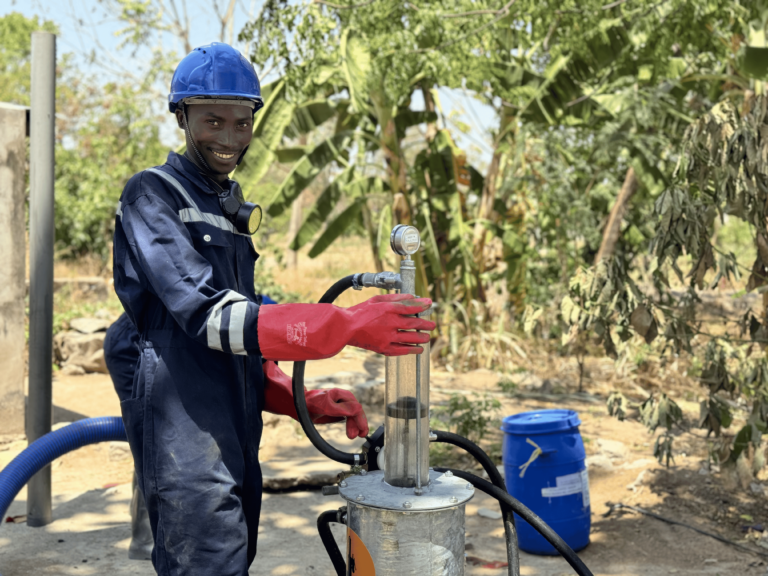 Man with blue overalls, red gloves and a blue hardhat stood smiling as he operates the PuPu pump. Lush green banana trees in the background