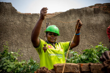 Naume dressed in green t-shirt and hard hat holding up a tool in front of a brick wall she is building